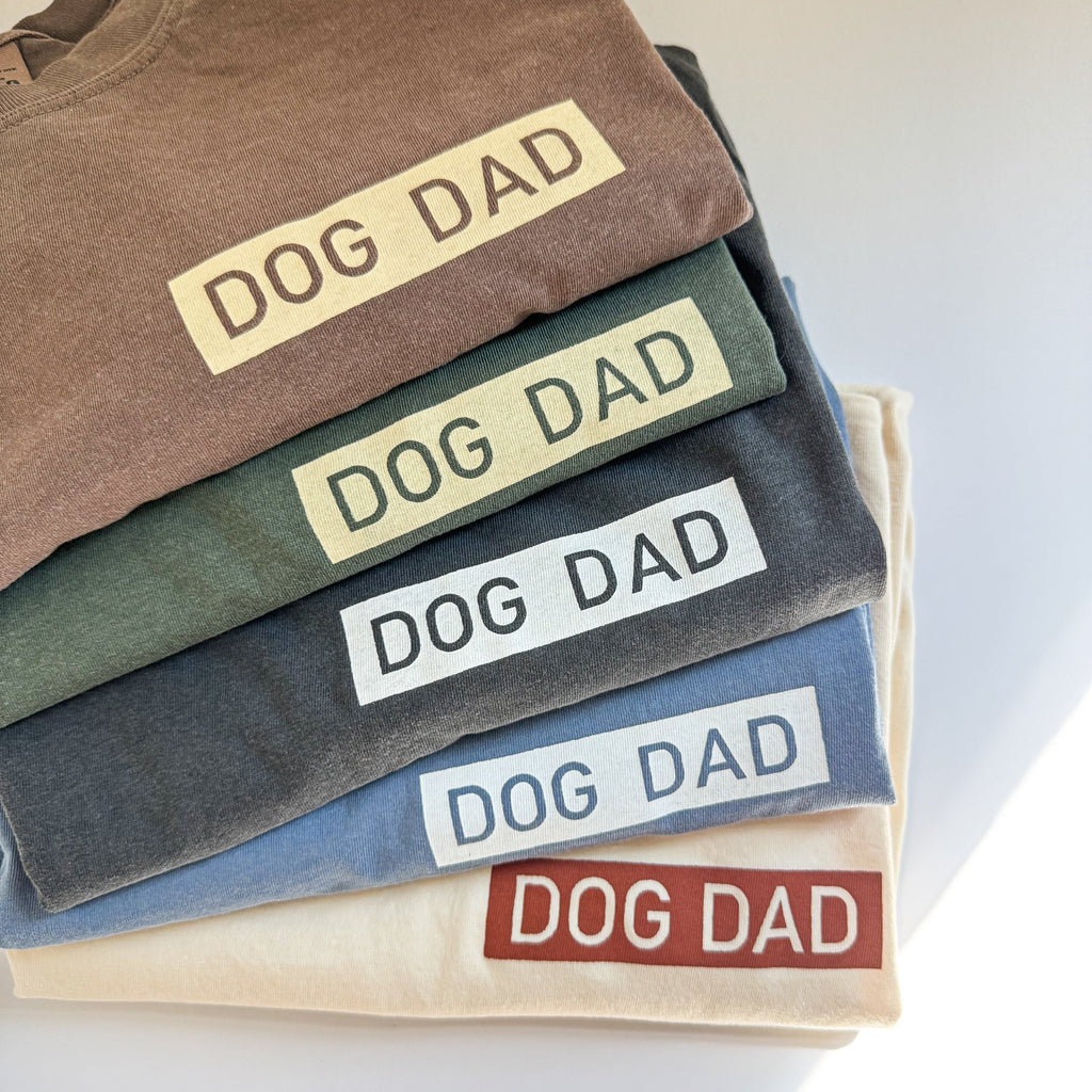 Stack of folded t-shirts with 'DOG DAD' text in various colors on a light background